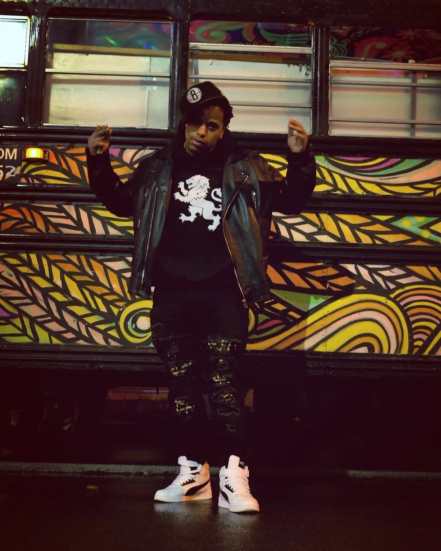 Kaleb Simmonds standing in front of a painted school bus wearing a leather jacket and a lion crest shirt